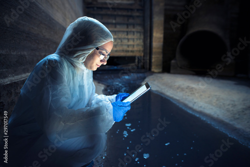 Ecologist expert or biotechnologist inside sewage sending report of wastewater contamination via tablet computer. Examining polluted water coming from sewage system into the river.