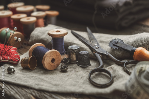 Vintage sewing items: retro tailoring scissors, measuring tape, thimble, wooden spools of thread, cutting knife, cushion for including pins, needles and sewing accessories.