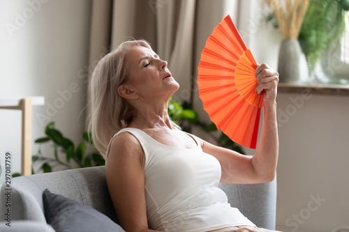 Stressed old woman waving fan suffer from overheating at home