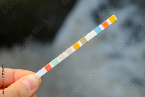 Strip for water quality measurement held in fingers (female hand). Ten multicolor squares for chemical testing of different reagents in water to identify its purity. Background of waterfall.