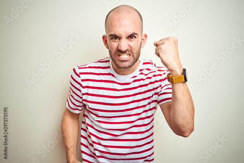 Young bald man with beard wearing casual striped red t-shirt over white isolated background angry and mad raising fist frustrated and furious while shouting with anger. Rage and aggressive concept.