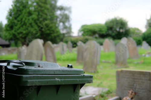 Green waste wheelie bin seen within a church's cemetery, which is used for discarded flowers.