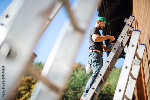 A side view of mature man outdoors in summer, painting wooden house.