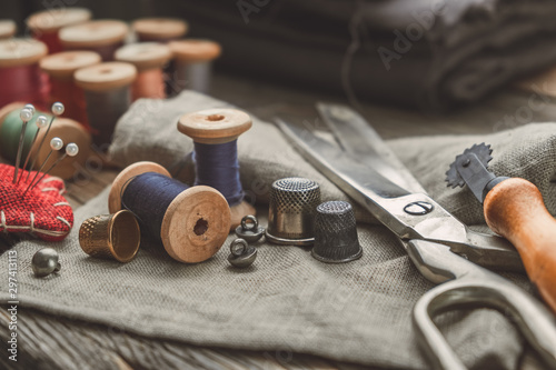 Retro sewing items: tailoring scissors, cutting knife, thimble, wooden thread spools, cushion for including pins, fabrics and sewing accessories.