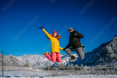 Young cheerful couple in bright clothes jump on the background of snowy mountains on winter sunny day. Guy and girl have fun during new year holidays in the mountains. Christmas and winter activities.
