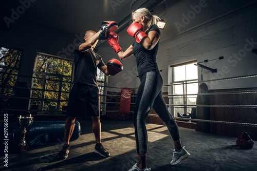 In the dark gym on the ring experienced trainer and young woman has a kick boxing fight.