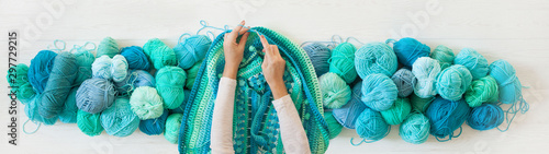 Women's hands are large. Woman crochets. Yarn of green, turquoise, aquamarine and blue colors. White wood long background.