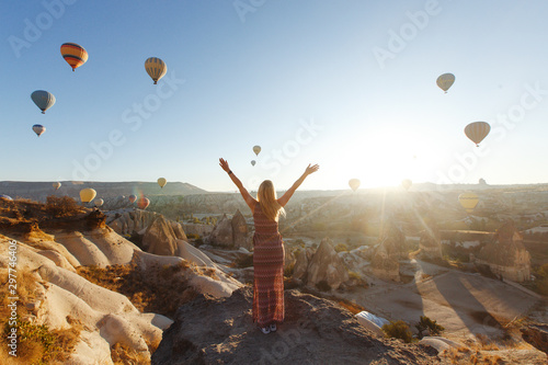 Young attractive girl in a hat stands on the mountain with flying air balloons on the background.Finger pointing girl in the sunrise. View from the back.Famous tourist Turkish region cappadocia.Gorem.