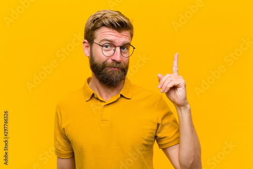 young blonde man feeling like a genius holding finger proudly up in the air after realizing a great idea, saying eureka against orange wall