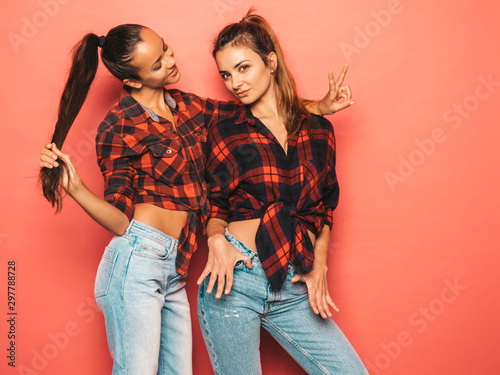 Two young beautiful smiling brunette hipster girls in trendy similar checkered shirt and jeans clothes.Sexy carefree women posing near pink wall in studio.Positive models having fun