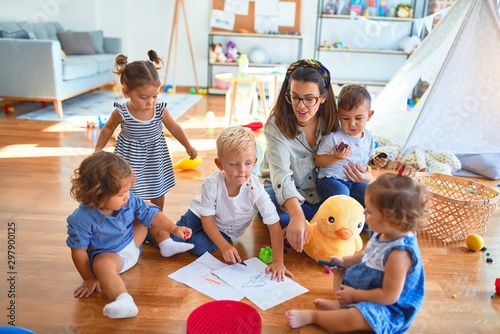 Beautiful teacher and group of toddlers sitting on the floor drawing using paper and pencil around lots of toys at kindergarten