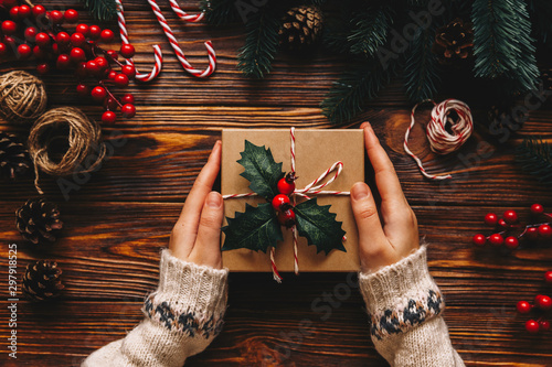 Christmas gift. Congratulations.Top wiew of woman holding traditional decorated gift box. Wooden table with cane,fir branches,fir cone, and berries