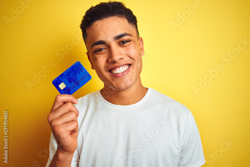 Young brazilian customer man holding credit card standing over isolated yellow background with a happy face standing and smiling with a confident smile showing teeth