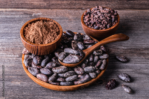 Whole dry cocoa beans, powder, nimbs in wooden bowls on old rustic background.