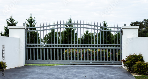 Grey metal wrought iron driveway property entrance gates set in white concrete brick fence, garden trees in background