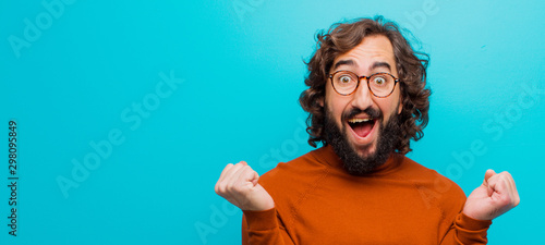 young bearded crazy man feeling shocked, excited and happy, laughing and celebrating success, saying wow! against flat color wall