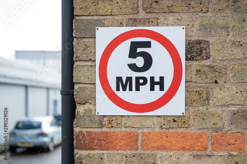 Detail of a 5MPH sign seen attached to a brick wall. The sign, located in a small industrial area, is to promote safety. A car can be seen parked in the distance.