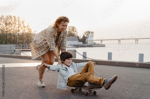 Young mother and son having fun with skateboard at the street. Happy family spending leisure time skateboarding at sunset.