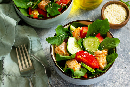 Tasty and healthy Salmon Grilled Salad with cucumber, tomatoes, fresh arugula and sesame seeds on a gray stone table.