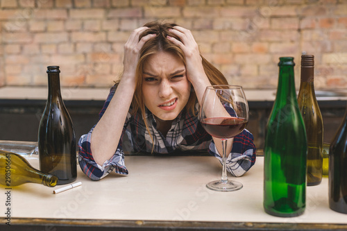 Young sad and wasted alcoholic woman sitting at kitchen couch drinking red wine, completely drunk looking depressed lonely and suffering hangover in alcoholism and alcohol abuse.