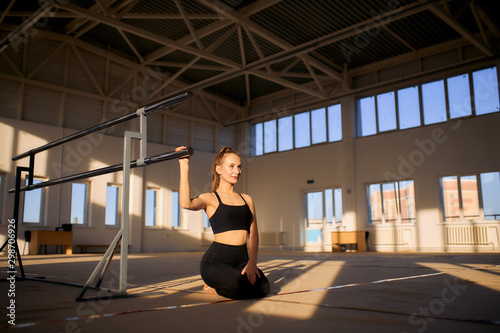 Beautiful charming girl with long hair, in black sportswear, sits on the floor in the middle of big gym hall, holds metal bar, looks away with positive smile, shot from below