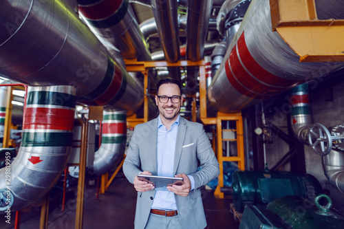 Handsome smiling supervisor in gray suit holding tablet while looking at camera. Energy plant interior.