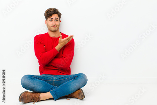 young handsome man feeling confused and clueless, wondering about a doubtful explanation or thought sitting on the floor