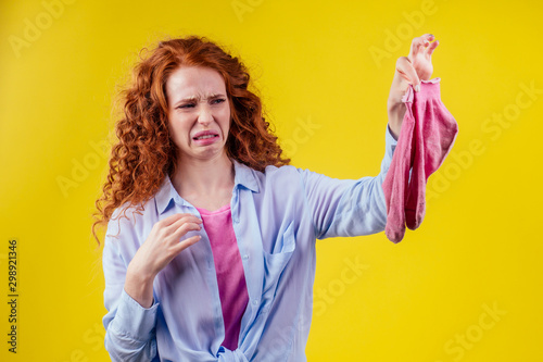 curly ginger redhead woman in a cotton shirt gesture smells bad holding a dirty pink sock out a disgusted look on her face in studio yellow background