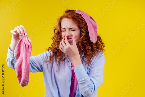curly ginger redhead woman in a cotton shirt gesture smells bad holding a dirty pink sock out a disgusted look on her face in studio yellow background