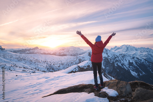 Happy young woman in snowy mountains at sunset in winter. Beautiful slim girl on the mountain peak with raised up arms, snow covered rocks and colorful sky with clouds. Travel in Dolomites. Tourism