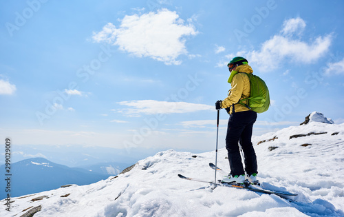Back view of sportsman skier in helmet and goggles with backpack standing on skis holding ski poles in deep white snow, on copy space background of bright blue sky enjoying beautiful mountain view.