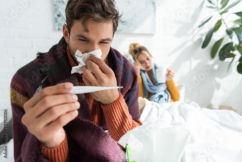 sick man with fever holding thermometer and napkin in bedroom with woman behind