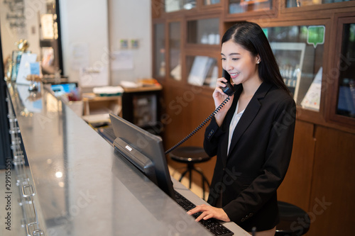 Welcome to the hotel,Happy young Asian woman hotel receptionist worker smiling standing,she taking telephone call at a Modern luxury reception counter waiting for guests getting key card in hotel