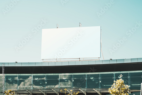 An empty huge poster mockup on the roof of a mall; white template placeholder of an advertising billboard on the rooftop of a modern building with glass facade; blank mock-up of an outdoor info banner