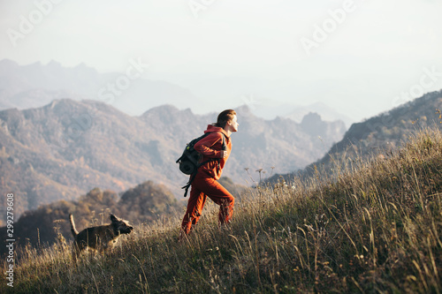 Beautiful woman traveler climbs uphill with a dog on a background of mountain views. She is with a backpack and in red clothes.