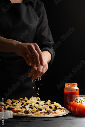 Chef cooks Italian pizza, sprinkles with mazarella cheese. Freeze in motion. Against the background of pizza ingredients. Black background, pizzeria, recipe book,restaurant business. Vertical photo.