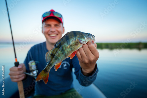 Happy amateur angler holds perch fish (Perca fluviatilis) in one hand and fishing rod in the other. Fisherman showing the fish and smiles being on the lake