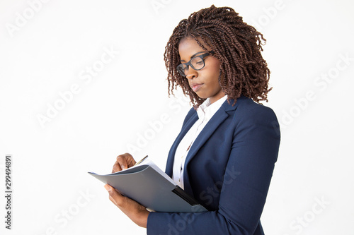 Focused accountant taking notes in paper, holding pen and folder, checking document. Young African American business woman posing isolated over white background. Paperwork concept