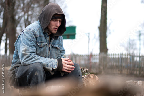 Homeless, sad man in a beanie sitting outside and feeling cold, trying to warm up his hands