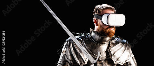 panoramic shot of knight with virtual reality headset in armor holding sword isolated on black