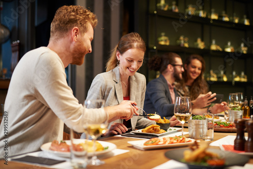 Handsome caucasian ginger taking food out of his girlfriend's plate while sitting in restaurant for dinner. In background are their friends.