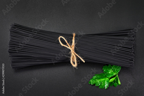 Bundle of black long raw spaghetti with green leaf of parsley on black background. Top view.