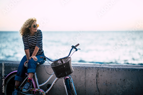 Happy beautiful curly healthy caucasian adult middle age woman sit down on a vintage coloured bike and enjoy the ocean in outdoor leisure activity - trendy people with hapy life