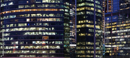 Glass walls of modern office buildings with many large panoramic windows in downtown business cluster with working people panoramic front view at night