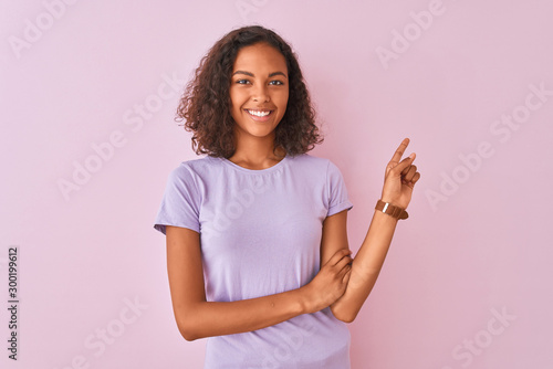 Young brazilian woman wearing t-shirt standing over isolated pink background with a big smile on face, pointing with hand finger to the side looking at the camera.