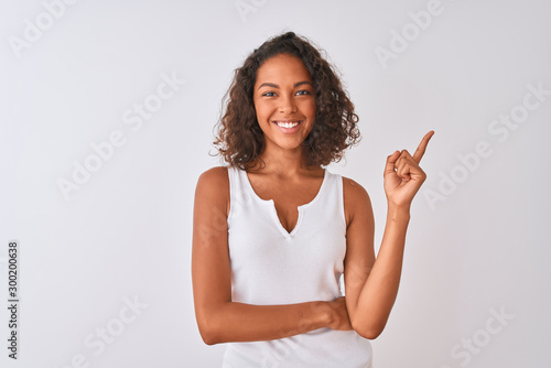 Young brazilian woman wearing casual t-shirt standing over isolated white background with a big smile on face, pointing with hand and finger to the side looking at the camera.