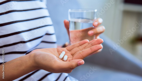 Woman holding white pill on hand and drinking water in glass on sofa in house, feels like sick. Health care concept.