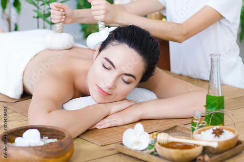 Young pleased woman is getting thai massage, therapy. Female master is massaging client with herbal bags. Brunette girl is lying on couch in light spa ayurveda salon. Relax and health care concept.