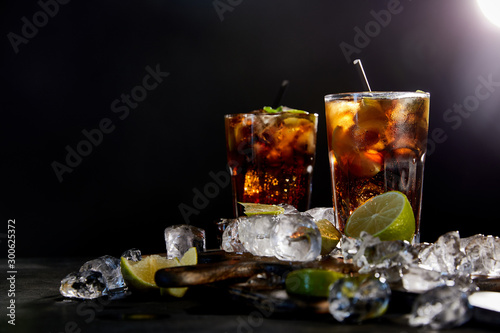 cocktails cuba libre in glasses with straws, ice cubes and limes on black background