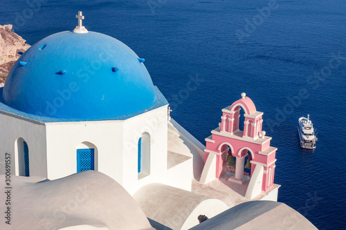 Blue roofed church with pink bell construction and yacht on the sea in Santorini, Greece. Travel, religion, yachting, boat, marine, culture concept.